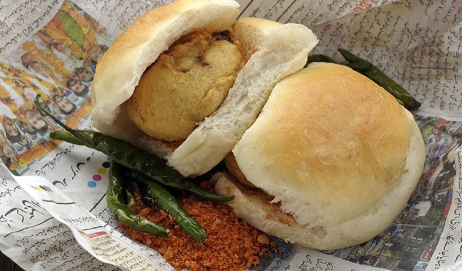 A Mumbai vada pav on newspaper with fried green chillies and dry garlic chutney